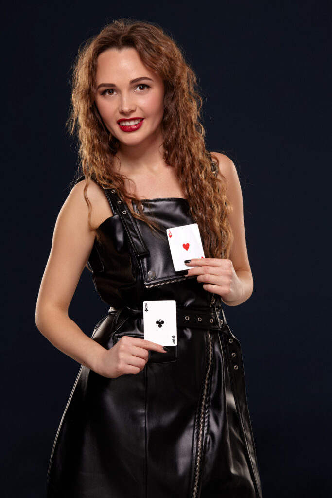 Emotional pretty young redhead woman holding pair of aces in black leather dress on black background